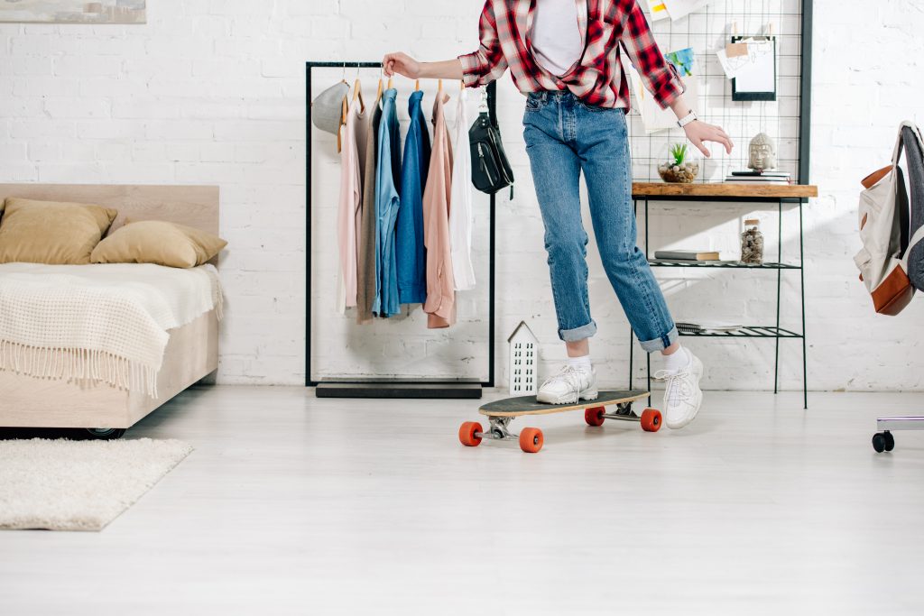 Cropped view of teenager in jeans and checkered shirt standing on longboard in bedroom
