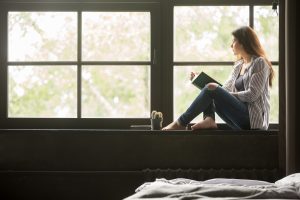 Pensive dreamy girl holding book sitting on sill at home looking at big window dreaming, thoughtful young woman thinking of reading new novel relaxing in modern cozy loft bedroom alone